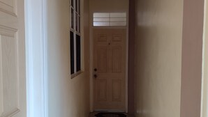 Un corridoio stretto con una porta in legno in fondo, una finestra con persiane e un lampadario.