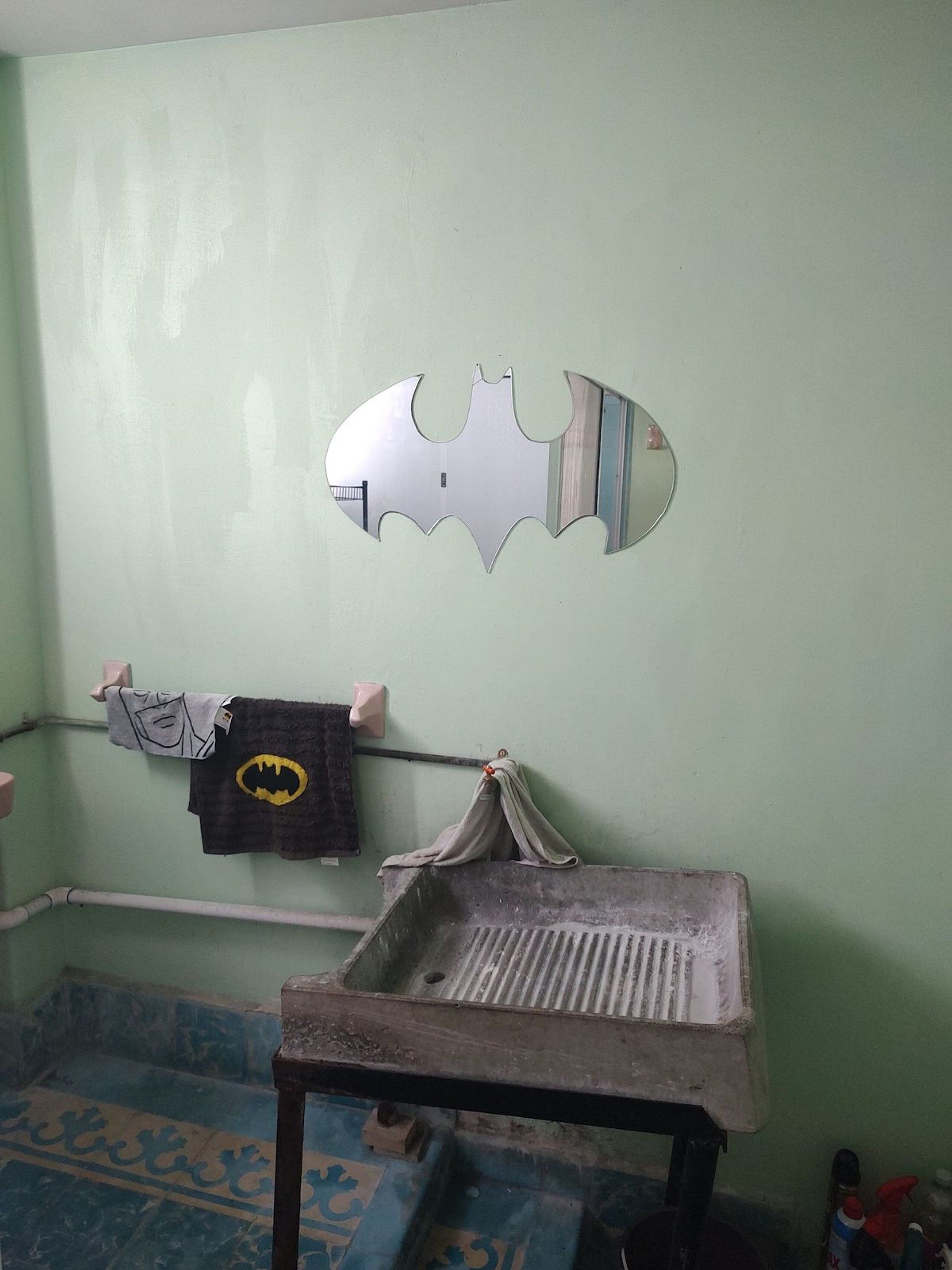 Un baño con una pared verde, un espejo con el logo de Batman, un perchero con una toalla de Batman y un lavabo con tabla de desagüe.