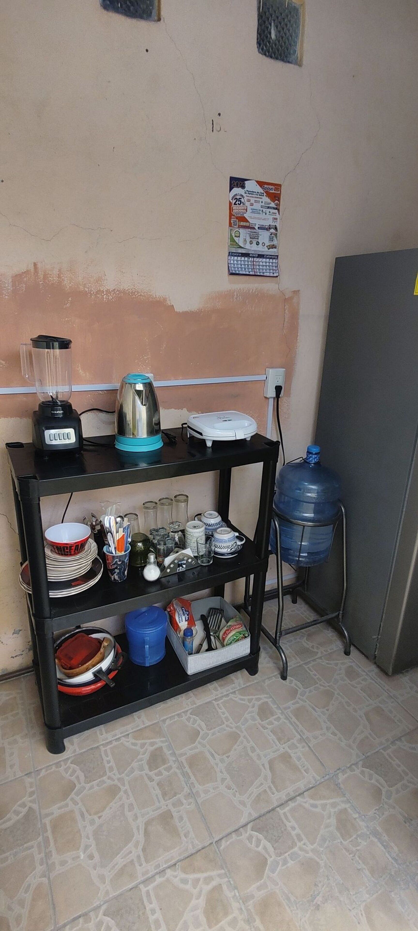 A kitchen area with a black shelf holding various kitchen items, a kettle, a blender, and a water dispenser.