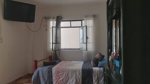 Un dormitorio con cama, ventana con cortinas, televisor montado en la pared y armario.