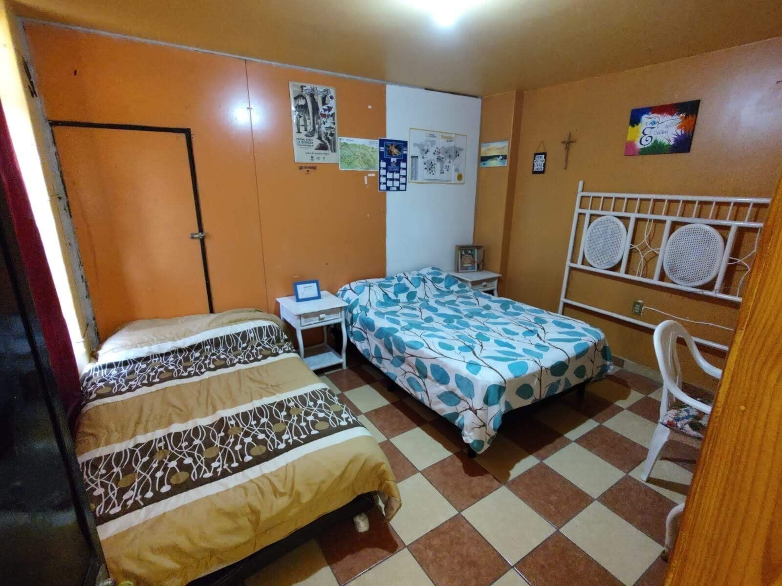 A bedroom with two beds, a desk, and a chair. There are posters on the wall and a window with bars.