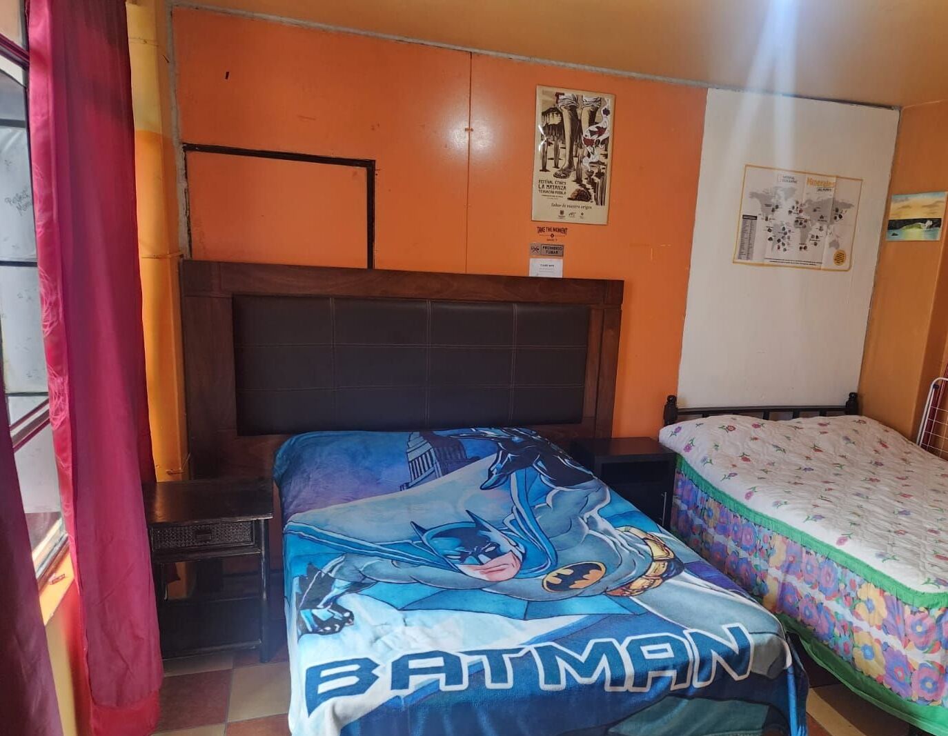 A room with two beds, one with a Batman-themed bedspread and another with a floral bedspread. There is a wooden headboard, a wall with posters, and a window with a red curtain.