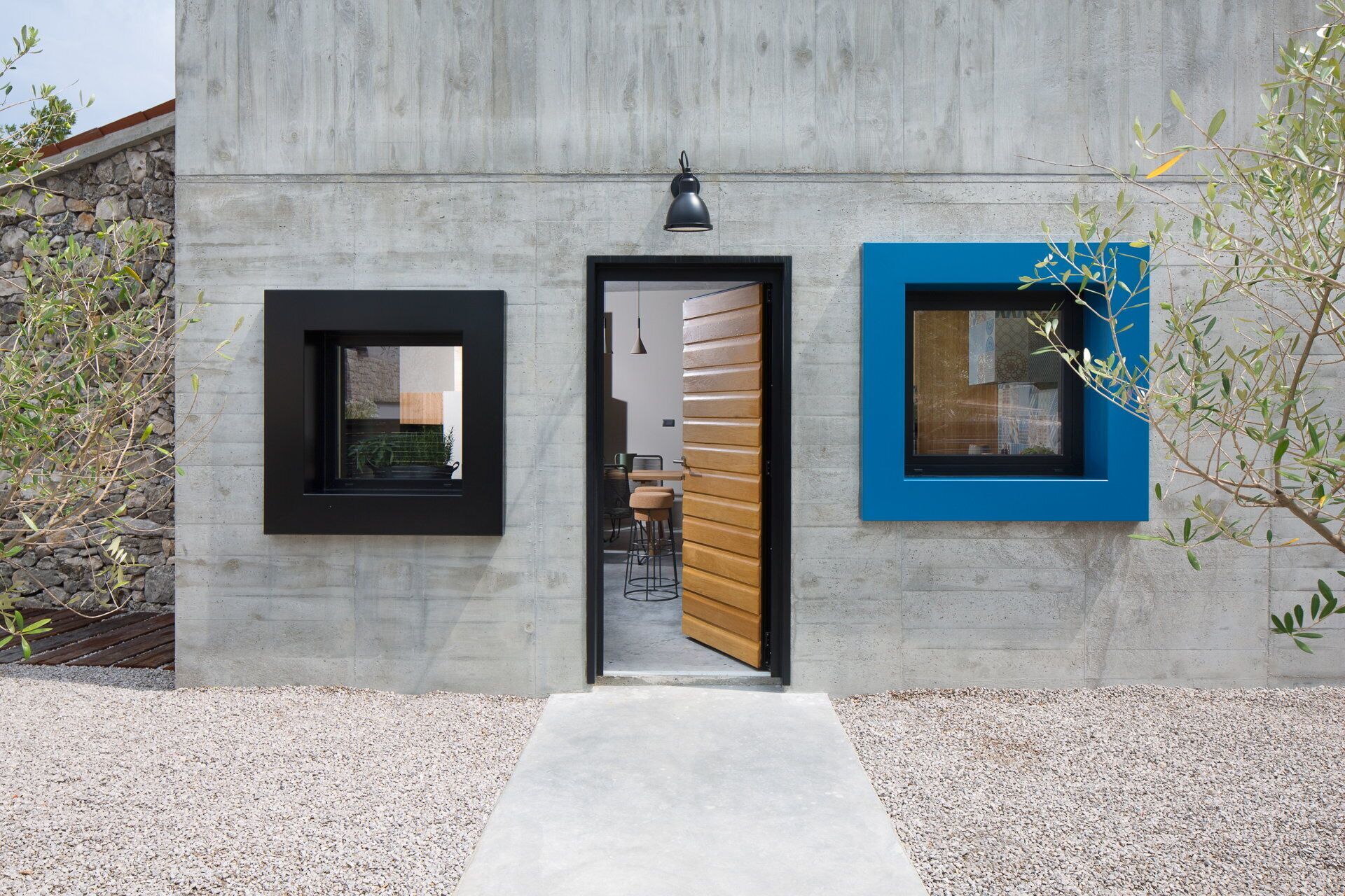 Un bâtiment moderne doté d’une façade en béton, d’une porte en bois et de deux fenêtres aux encadrements noirs et bleus.