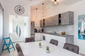 A modern kitchen with grey cabinets, a white countertop, and a dining area with a white table and grey chairs. There are two pendant lights, a clock on the wall, and a painting of a sailboat on the wall. - Villa Samba with Pool (Pula)
