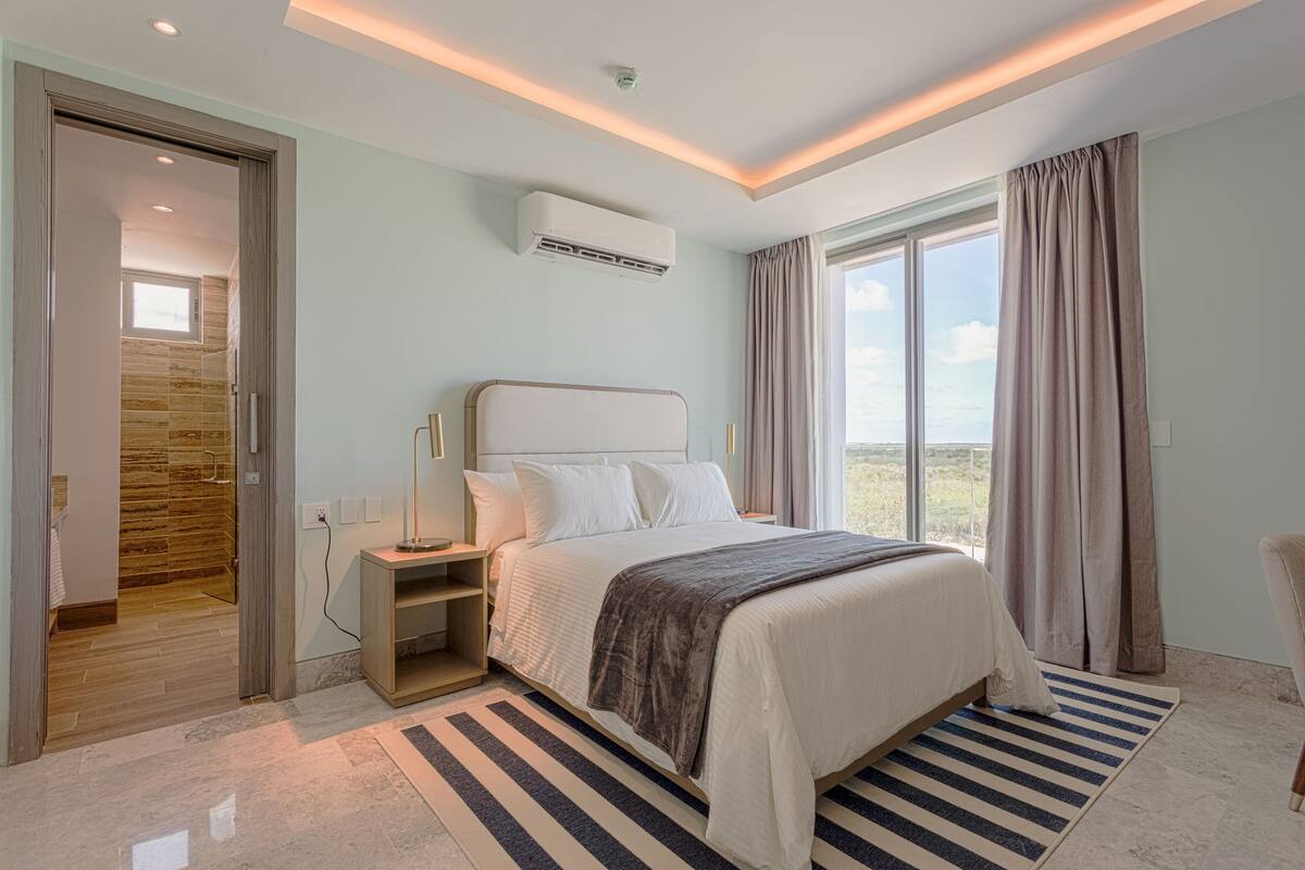 A modern bedroom with a large bed, a nightstand, a wall-mounted air conditioner, and a view of the outdoors through a window with curtains.
