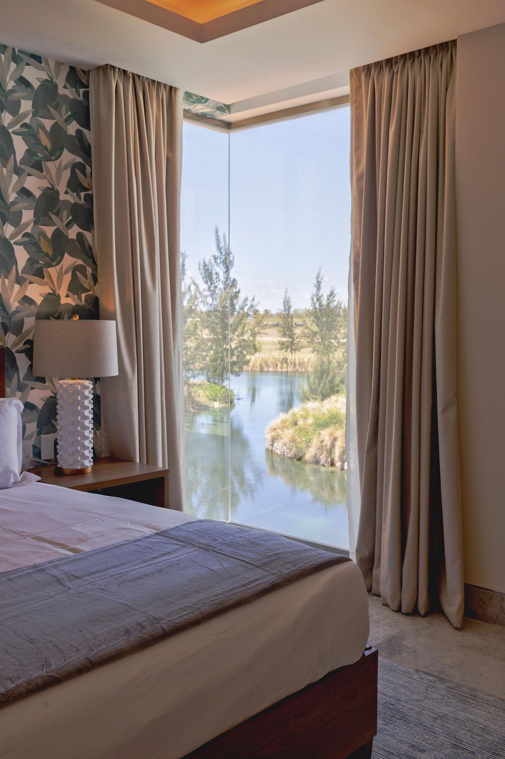A bedroom with a large bed, a bedside table with a lamp, and a view of a lake and trees through floor-to-ceiling windows.