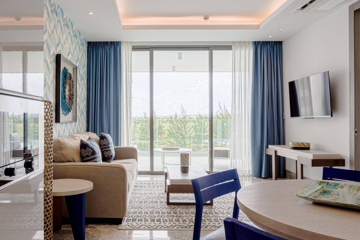 A modern living room with a sofa, a coffee table, and a TV. There is a large window with curtains, a patterned rug, and a view of greenery outside.