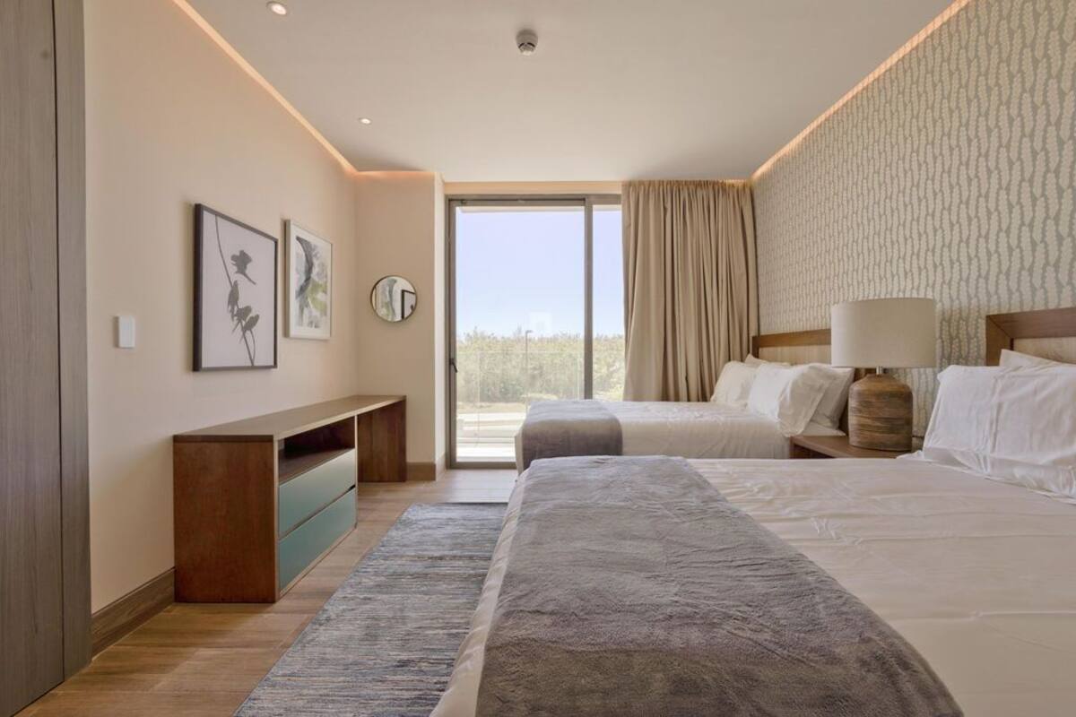 A modern hotel room with two beds, a large window, a view of the outdoors, a wooden bench, and a wall-mounted clock.
