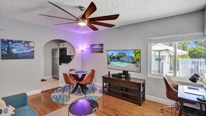 A modern living room with a ceiling fan, a dining area with orange chairs, a TV, and a view of a backyard with a pool. - Retro Oasis by Las Olas (Fort Lauderdale)