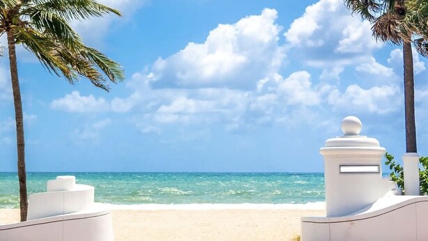 A beach with white structures, palm trees, and a clear blue sky. - Retro Oasis by Las Olas (Fort Lauderdale)