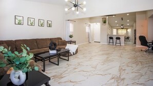 A spacious living area with a brown sofa, a coffee table, and a potted plant. There is a kitchen area in the background with a bar counter and stools.