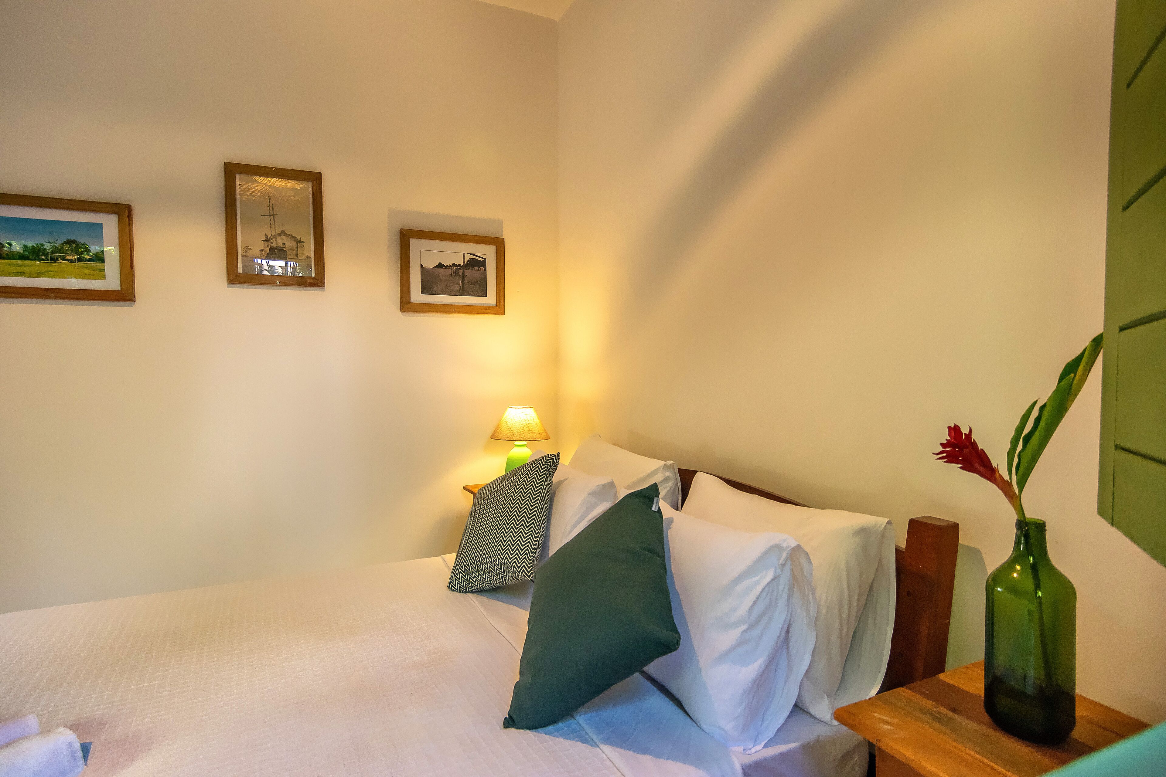 A neatly made bed with white and green pillows, a small bedside table with a green vase, and two framed pictures on the wall.