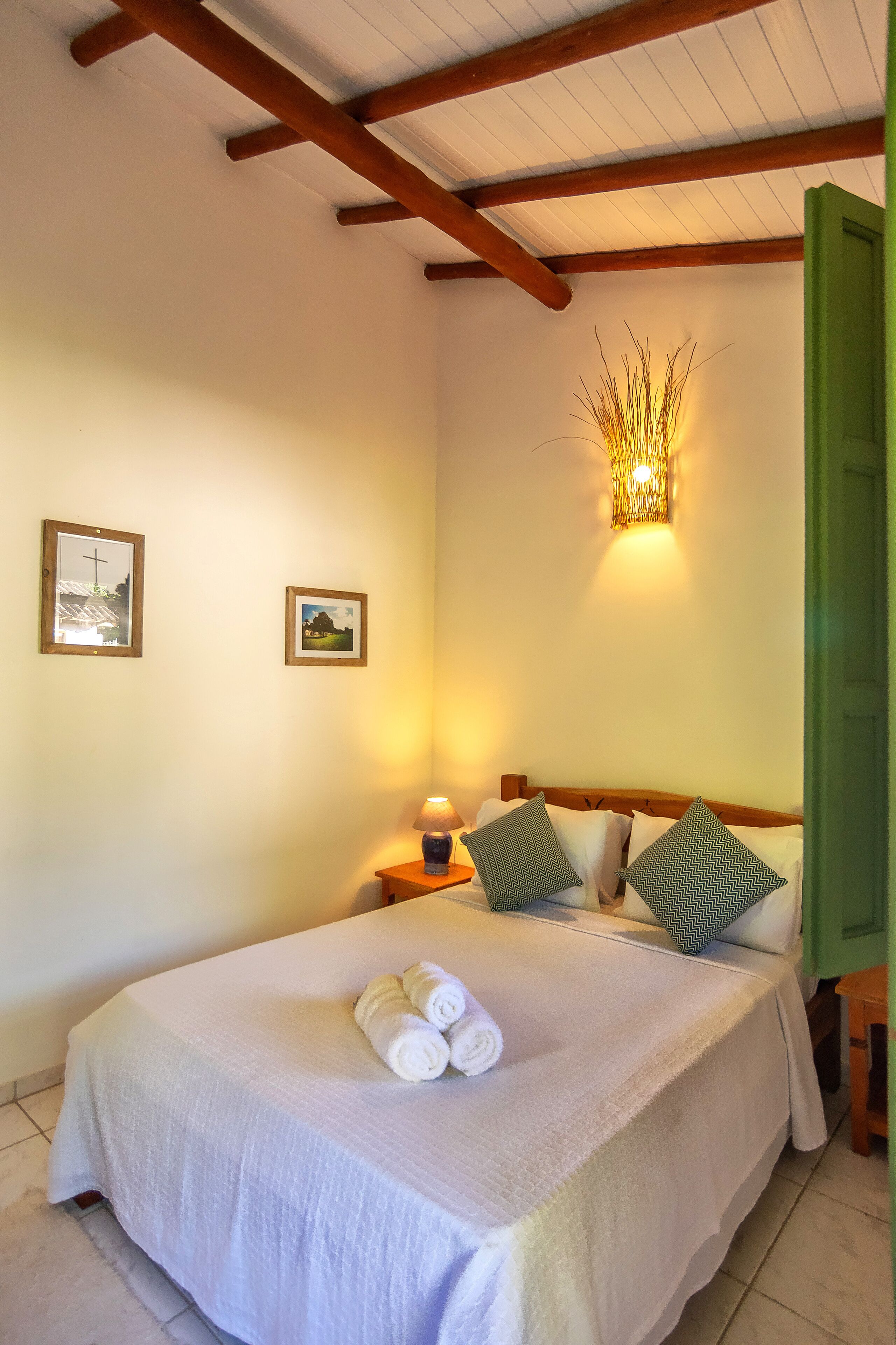 A neatly made bed with white linens and rolled towels, a bedside lamp, and two framed pictures on the wall.