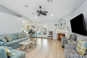 A living room with a sofa, coffee table, and patterned armchairs. There is a fireplace, a television, and a ceiling fan. - Family Oasis Stunning  Private Pool Home (Navarre)