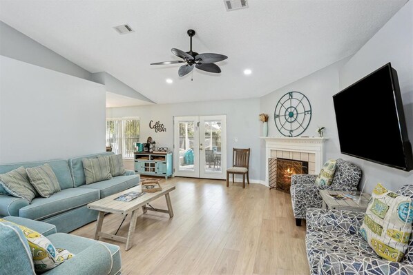 A living room with a sofa, coffee table, and patterned armchairs. There is a fireplace, a television, and a ceiling fan. - Family Oasis Stunning Private Pool Home (Navarre)