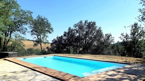 Una piscina rettangolare con bordo in legno, immersa in un giardino curato con alberi e uno sfondo collinare.