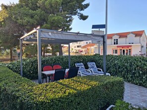 A covered outdoor seating area with tables and chairs, surrounded by neatly trimmed hedges and trees.