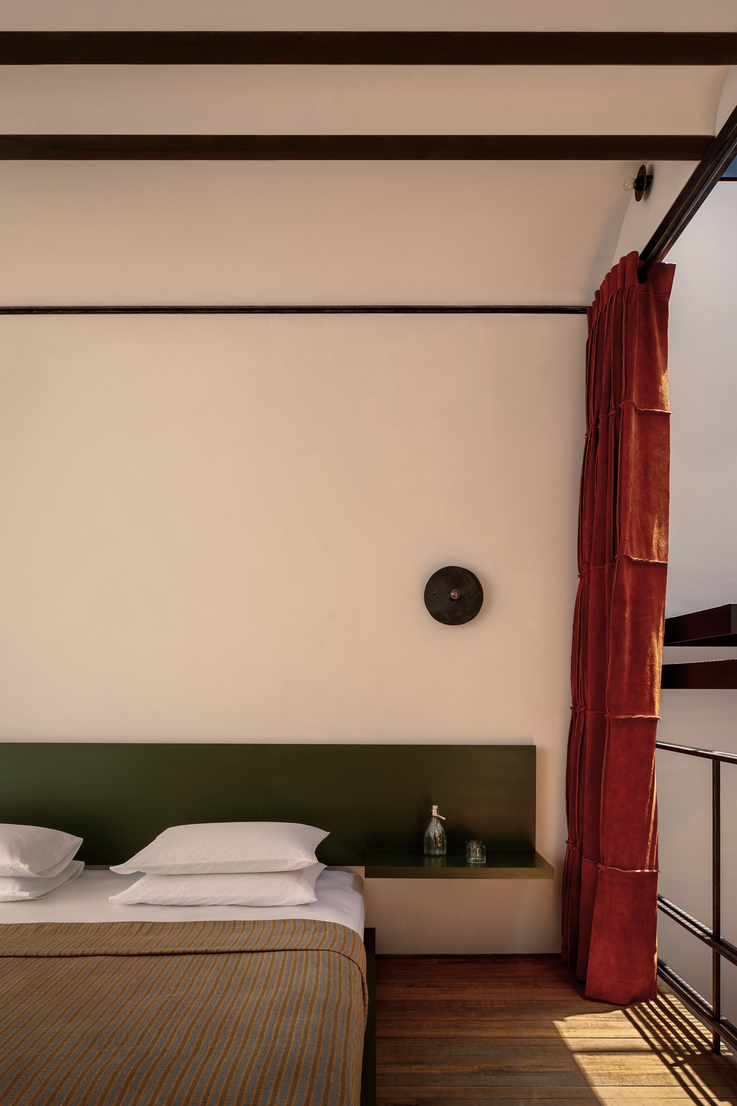 Un dormitorio con cama, almohadas, una mesita de noche, una cortina roja y un aplique de pared.