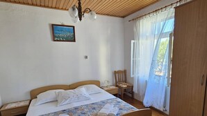 Une chambre à coucher avec un lit en bois, une table de chevet, une chaise, une armoire, un ventilateur de plafond et une fenêtre avec des rideaux.