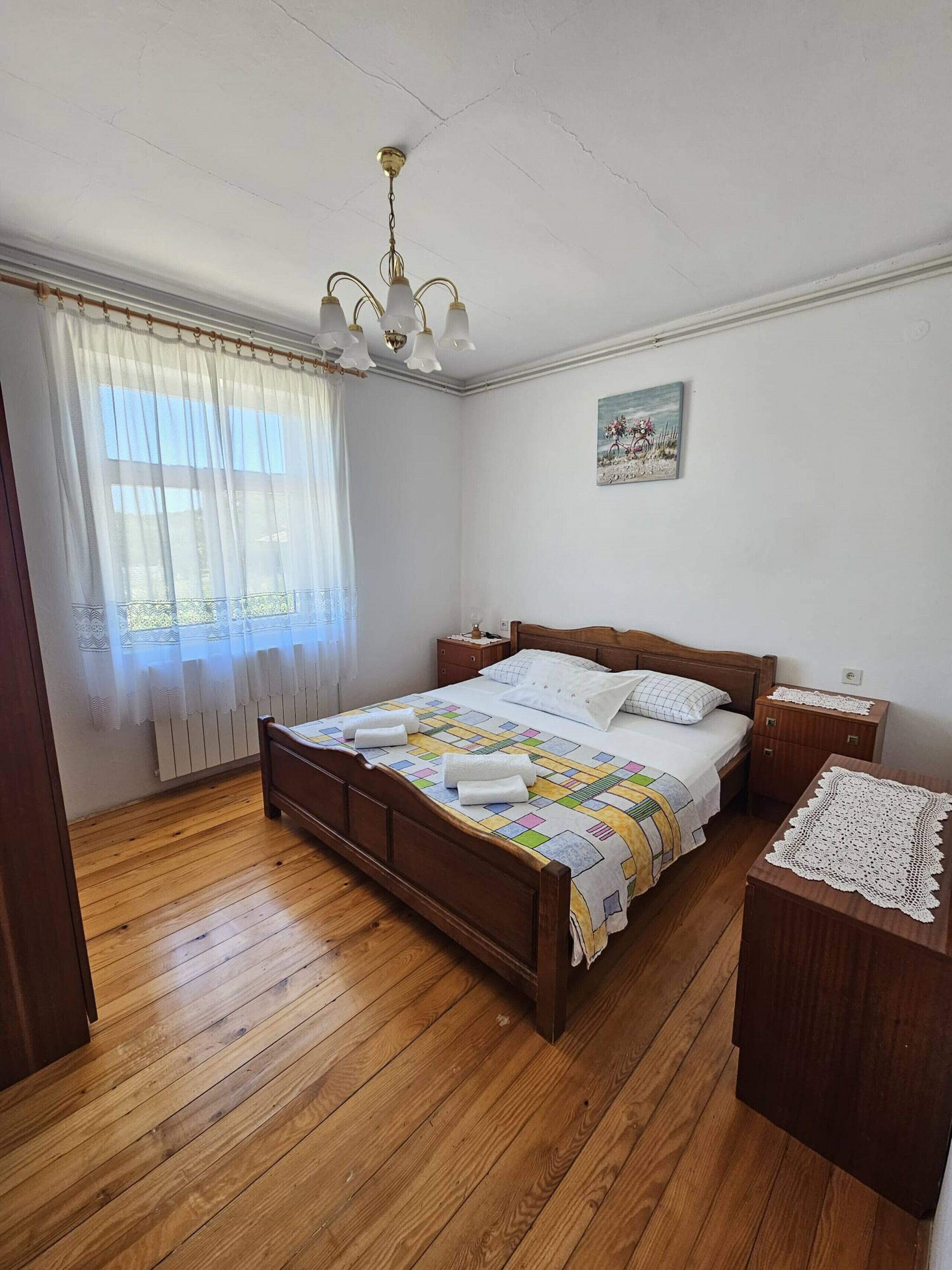 A bedroom with a wooden bed, white curtains, a ceiling light fixture, a bedside table, and a nightstand.