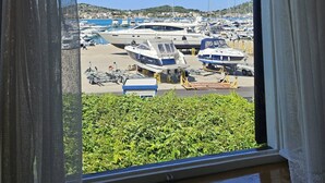 Une vue à travers une fenêtre ornée de rideaux blancs, dévoilant un port de plaisance où plusieurs bateaux sont amarrés, et un paysage verdoyant au premier plan.