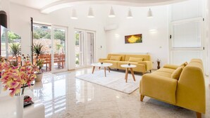 A spacious living room with a yellow sofa, two white coffee tables, and a large window with a view of greenery.