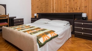 Un dormitorio con cabecera de madera, cama con ropa de cama blanca, mesita de noche con lámpara y cómoda con cajones.