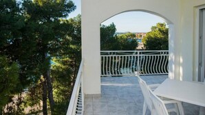 Un balcon avec une table et des chaises blanches, une rambarde en métal et une vue sur des arbres et un paysage lointain.