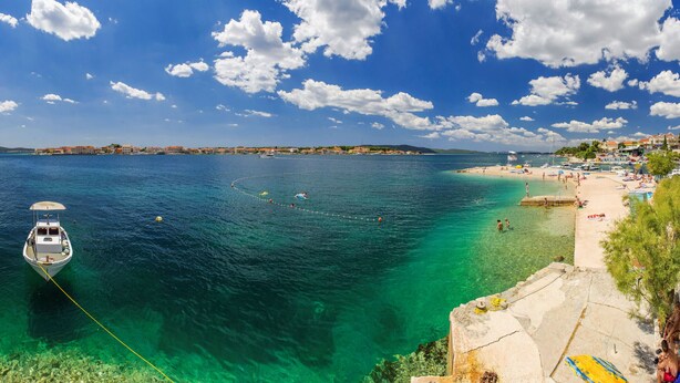 A coastal scene with a boat anchored near the shore, clear turquoise waters, and a beach with people and sun loungers. - Apartments Gaj Brodarica - A9 (Šibenik-Brodarica)