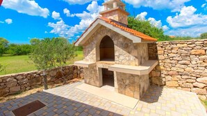 A stone-built outdoor kitchen with a chimney, surrounded by a stone wall and a paved patio.