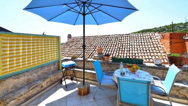 Un patio con una sombrilla azul, dos sillas, una mesa con frutas y un tejado de baldosas al fondo.