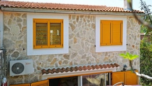 Un edificio de piedra de dos pisos con contraventanas amarillas, un techo de tejas y una unidad de aire acondicionado visible.