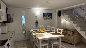 A dining area with a white table and chairs, a kitchen with white cabinets, a sofa with colorful cushions, and a staircase with metal railings. - Stone Apartment Primosten (Primošten)