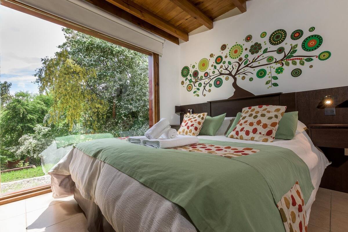 A bedroom with a large window, a bed with green bedding, a tree mural on the wall, and a view of greenery outside.