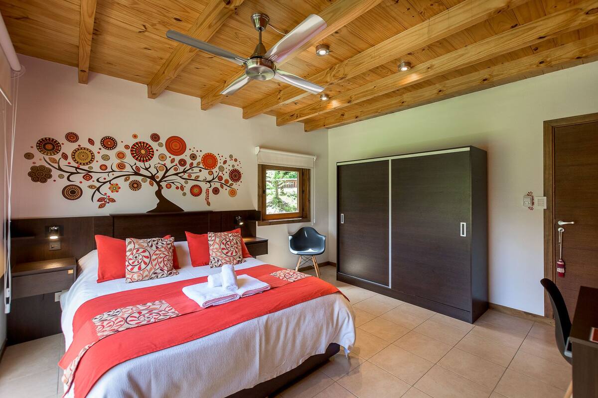 A hotel room with a wooden ceiling, a bed with red and white bedding, a wall mural, a wardrobe, and a window.