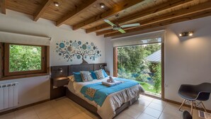 A bedroom with a large bed, wooden ceiling beams, a ceiling fan, a window with a view of greenery, and a chair by the window. - Solar Selvana - Casas de montaña (Villa La Angostura)