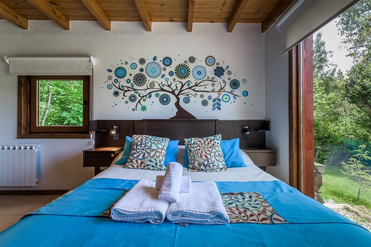 A bedroom with a large bed, a tree mural on the wall, and a view of greenery outside.
