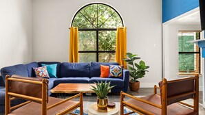 Un salon avec un canapé bleu, deux fauteuils en bois, une petite table et une grande fenêtre ornée de rideaux jaunes.