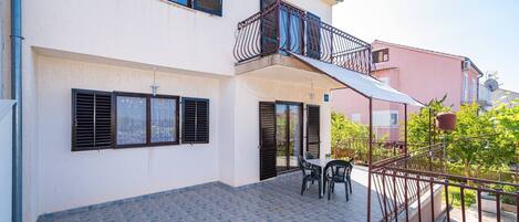 Edificio de dos pisos con patio empedrado, balcón con baranda de metal y una mesa con sillas.