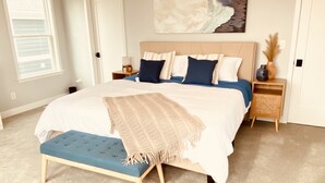 Un dormitorio con una cama grande, un banco, una mesita de noche, un cuadro en la pared y una puerta.