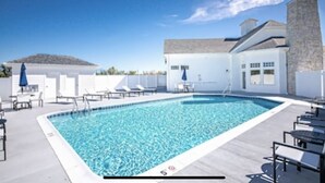 Una piscina rectangular con agua azul cristalina, rodeada de sillas de descanso y sombrillas, con una chimenea de piedra y un edificio blanco al fondo.