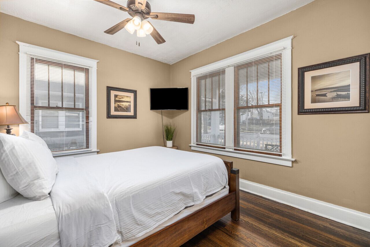 A bedroom with a bed, a ceiling fan, two windows with blinds, a wall-mounted TV, and framed pictures.