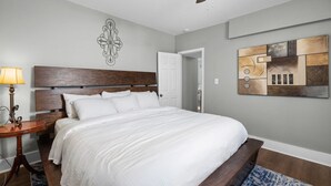 Een slaapkamer met een groot bed, een houten hoofdbord, een plafondventilator, een wanddecoratie met abstracte kunst en een nachtkastje met een lamp.