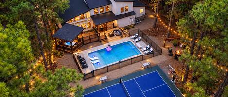 A two-story house with a swimming pool and tennis court, surrounded by trees.