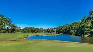 A golf course with a pond, surrounded by trees and a clear blue sky.