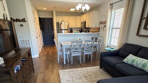 A modern living room with a dark blue sofa, a wooden coffee table, and a grey rug. A kitchen with white cabinets and a dining area with white chairs is visible in the background.