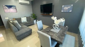 Un salón moderno con un sofá gris, una mesa de comedor con sillas, un televisor de pantalla plana montado en la pared y un cuadro decorativo de un elefante.