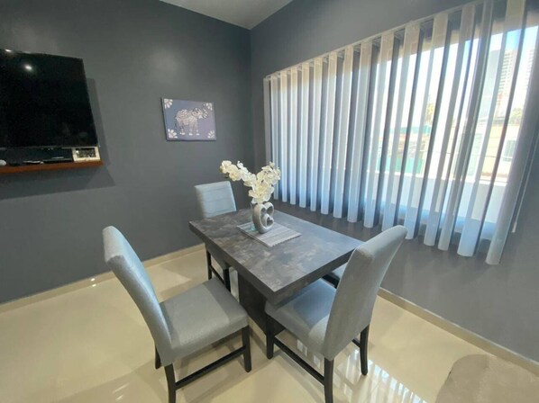 A dining area with a table, chairs, a vase with flowers, a flat-screen TV, and a window with blinds.