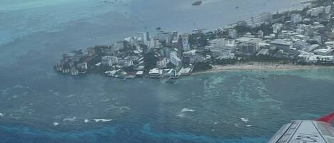 An aerial view of a coastal city with buildings near the shoreline, a clear blue sea, and a cloudy sky.