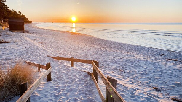 Ein Holzweg führt zu einem Strand, im Hintergrund ein Sonnenuntergang.